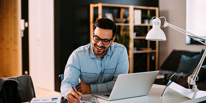 A young man working at home with a laptop and papers on his desk.
